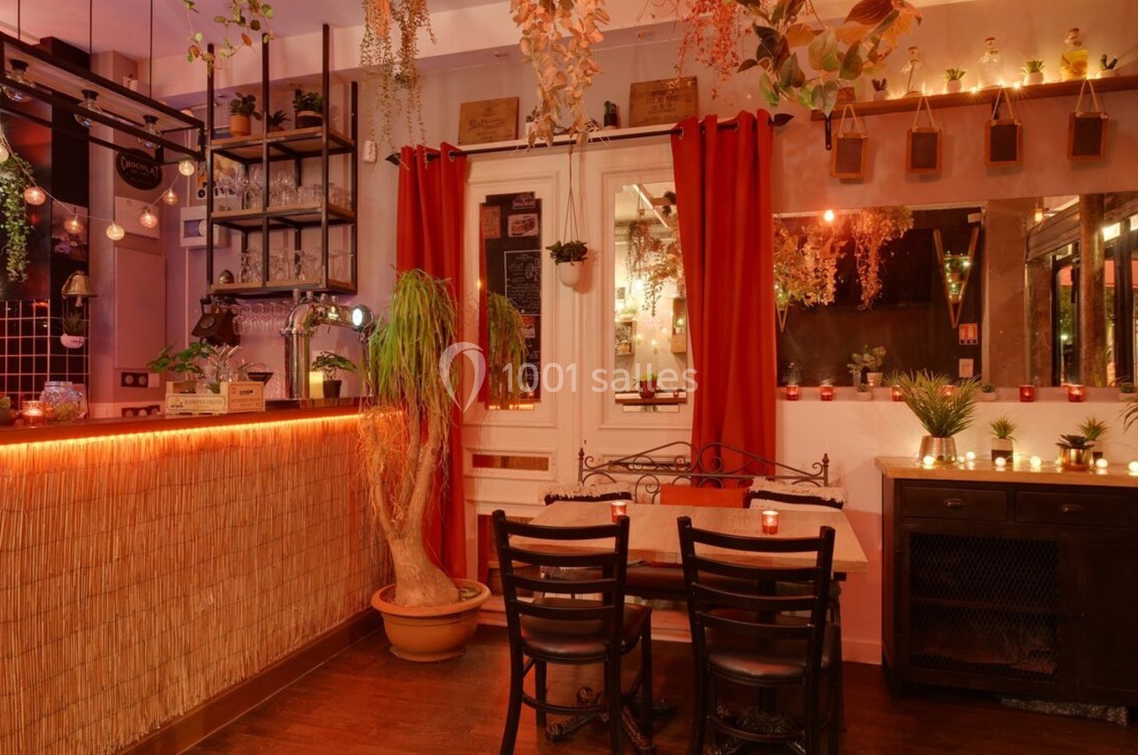 Intérieur d'un bar chaleureux avec éclairage tamisé, plantes suspendues et petite table entourée de chaises.