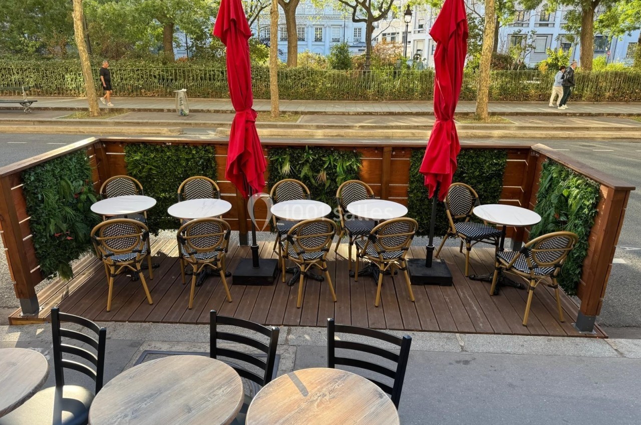 Terrasse en bois avec tables rondes, chaises en rotin et parasols rouges, bordée de plantes vertes, donnant sur une rue…