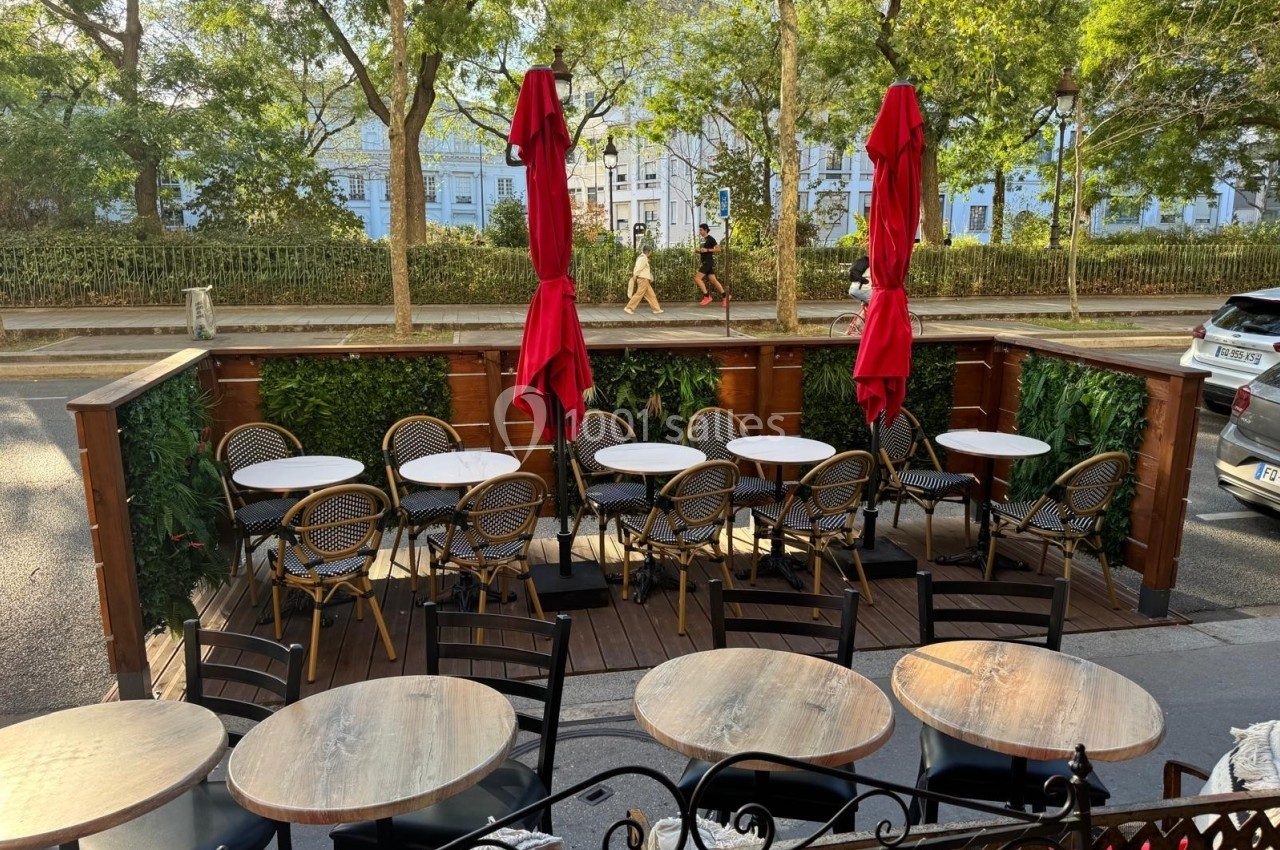 Terrasse extérieure avec tables rondes, chaises en rotin et parasols rouges, bordée de verdure et donnant sur une rue…