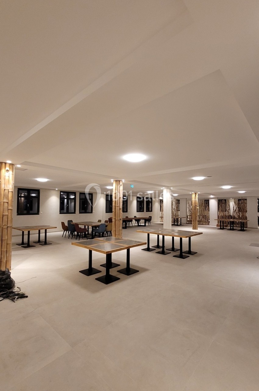 Salle spacieuse avec tables et chaises dispersées, éclairée par des plafonniers et entourée de colonnes décoratives.