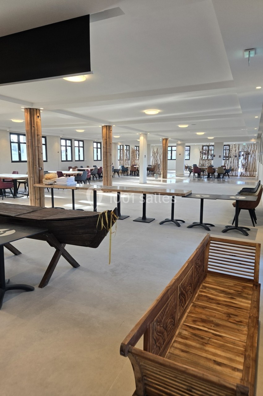 Salle spacieuse avec tables, chaises et bancs en bois, éclairée par des fenêtres et des luminaires au plafond.