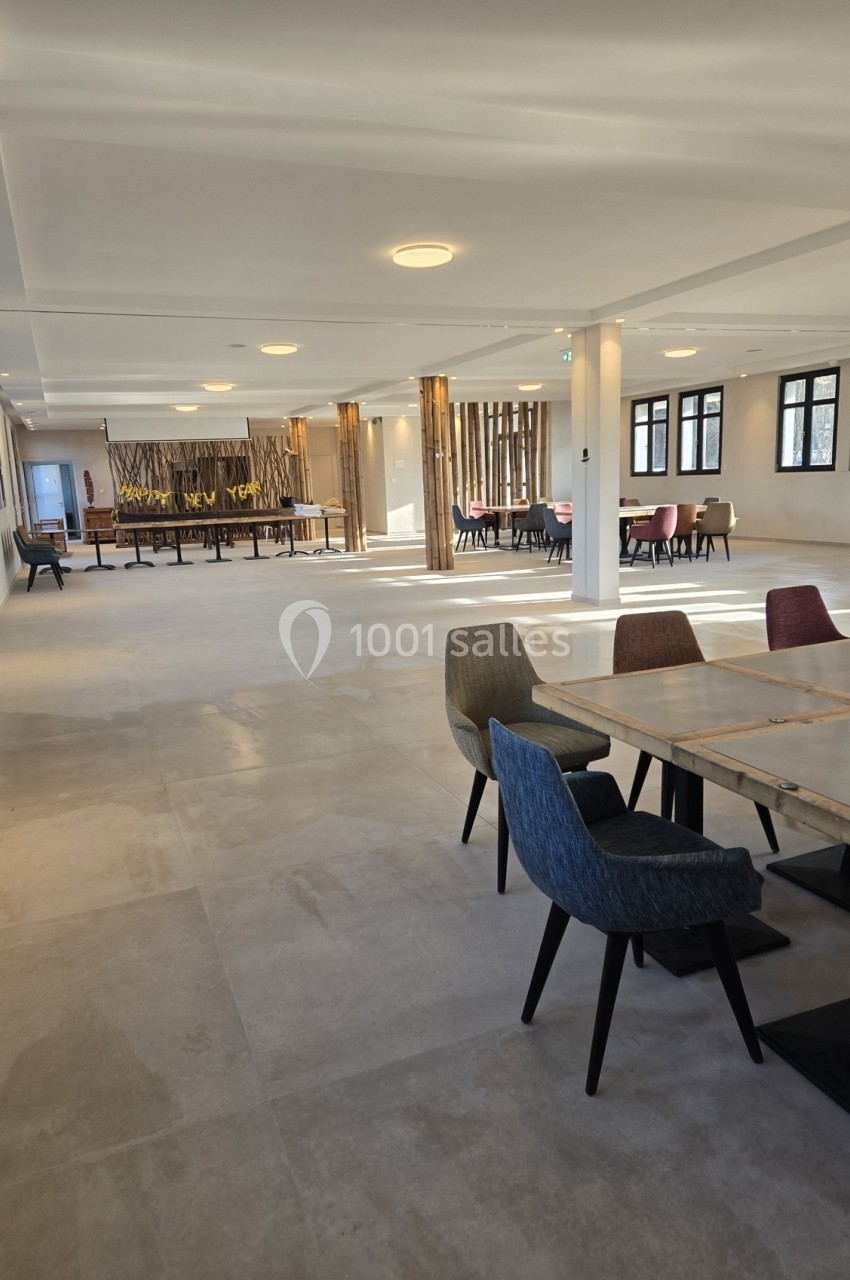Salle lumineuse avec tables, chaises colorées et grandes baies vitrées, décorée de panneaux en bois au fond.