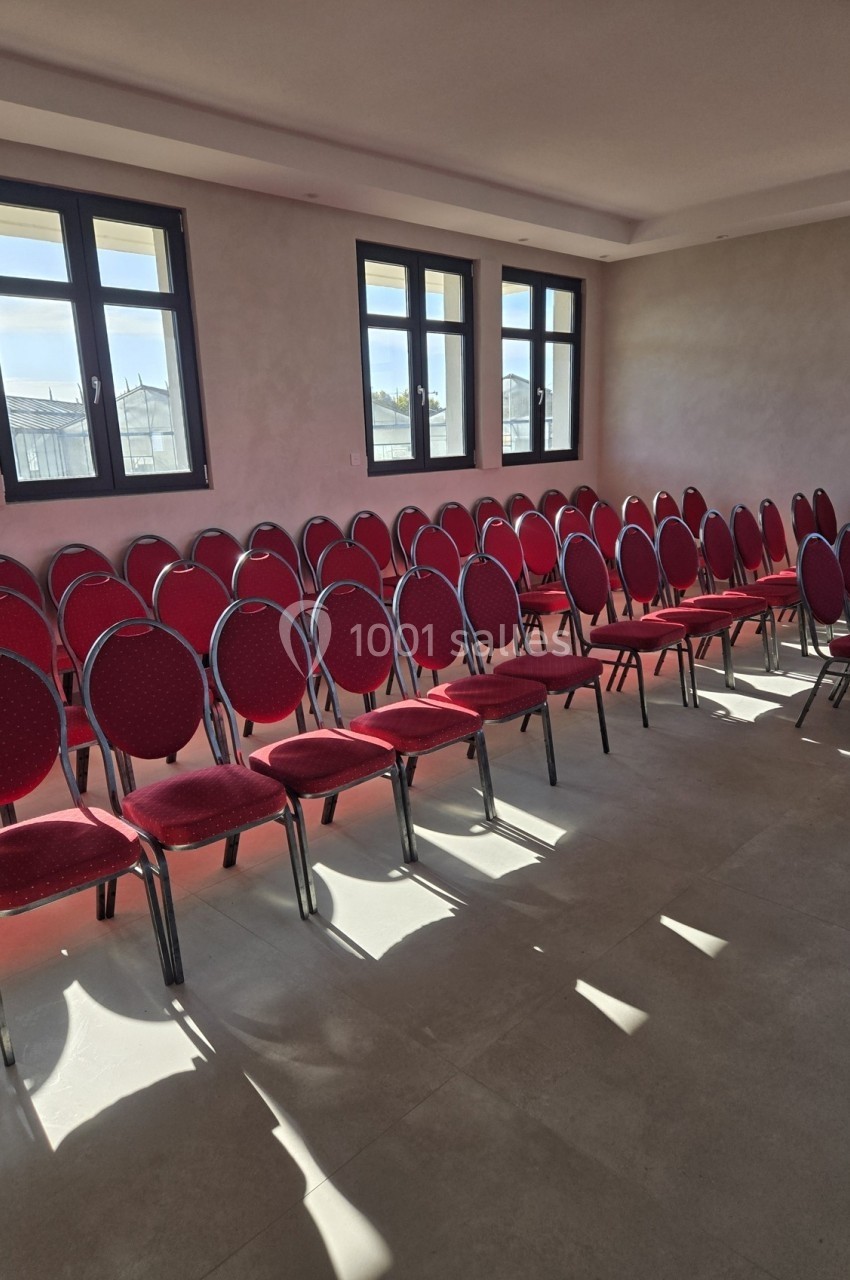 Salle lumineuse avec plusieurs rangées de chaises rouges alignées devant des fenêtres.