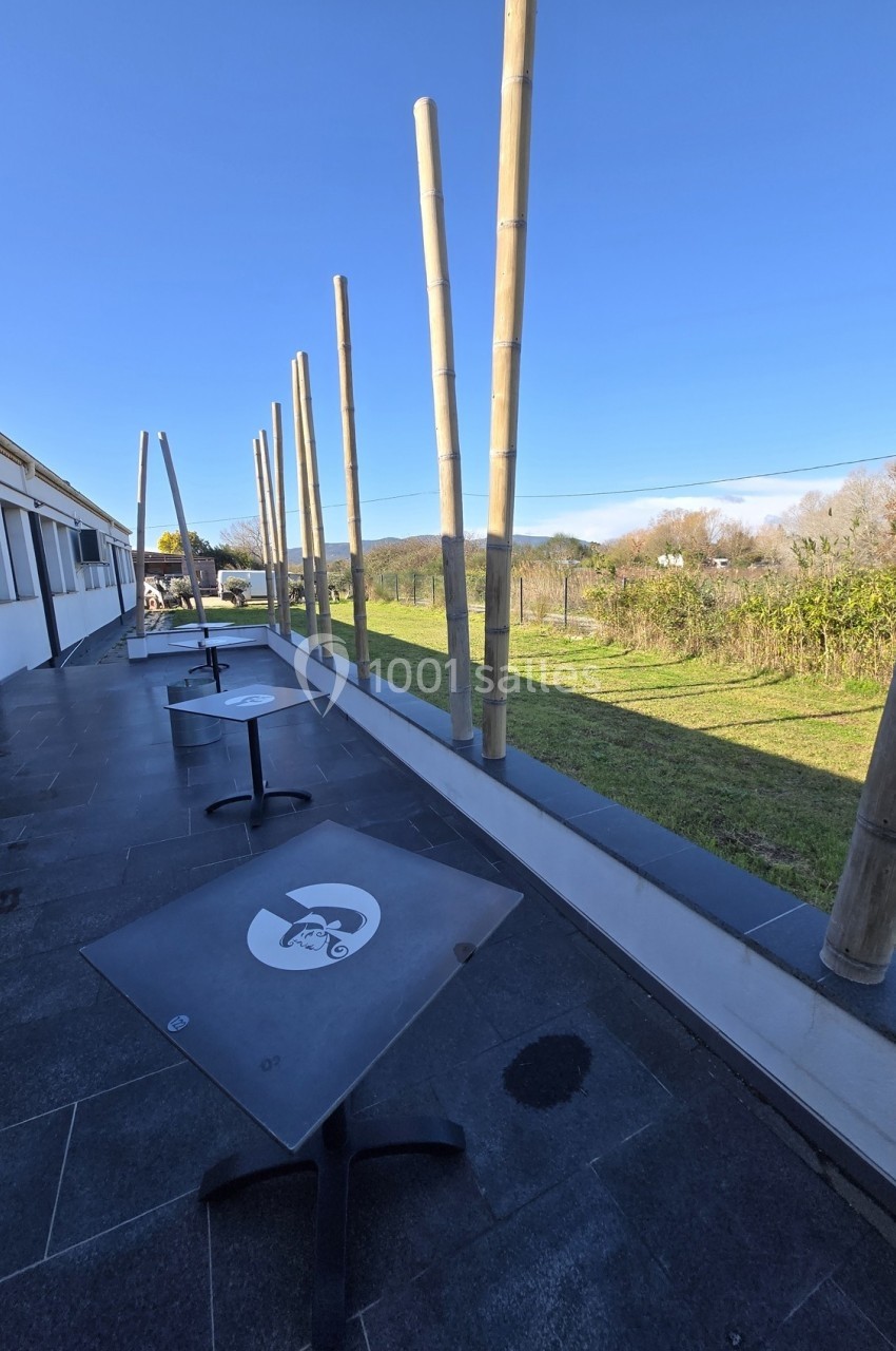 Terrasse avec tables noires et poteaux en bambou, offrant une vue dégagée sur un paysage verdoyant et ensoleillé.