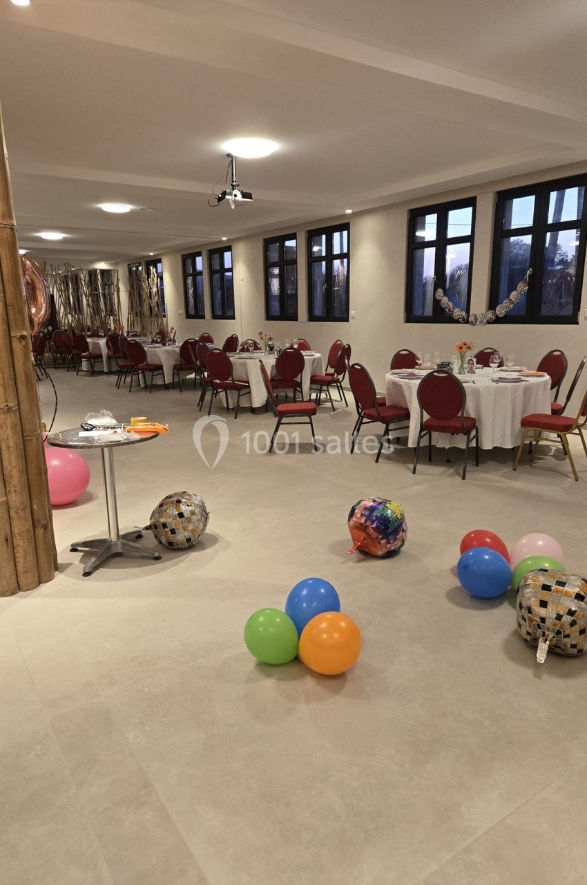 Salle décorée pour une fête avec des tables dressées, des ballons colorés au sol et des guirlandes en arrière-plan.