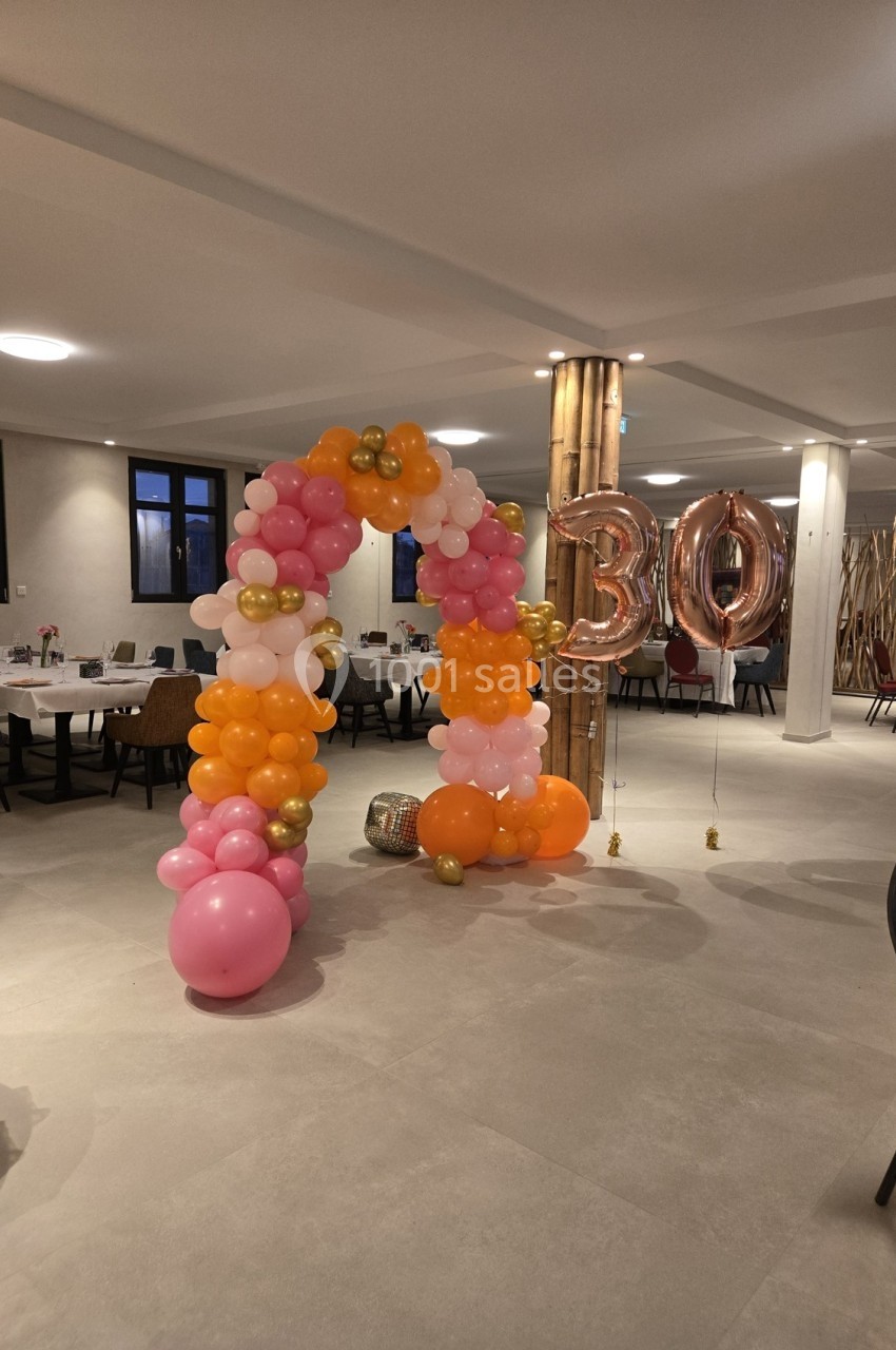 Arche de ballons roses, oranges et dorés avec des ballons ’30’ dans une salle de réception décorée.