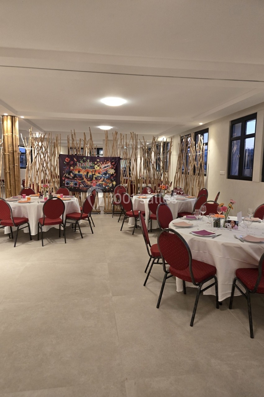 Salle de réception avec tables rondes dressées, chaises rouges et décorations en bambou devant une fresque colorée.