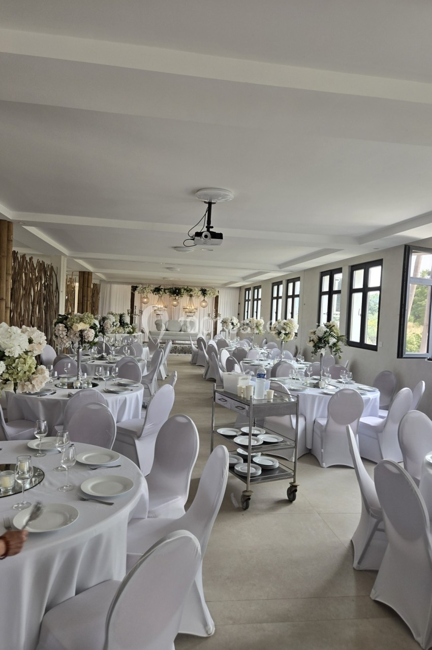 Salle de réception décorée avec des tables rondes, nappes blanches, chaises assorties et arrangements floraux élégants.