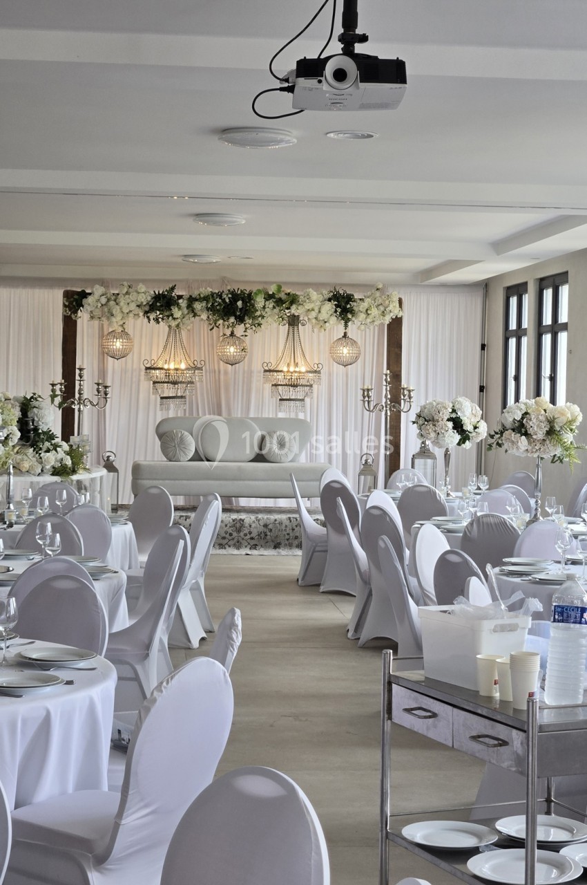 Salle de réception décorée pour un mariage, avec tables dressées et une scène ornée de fleurs blanches.