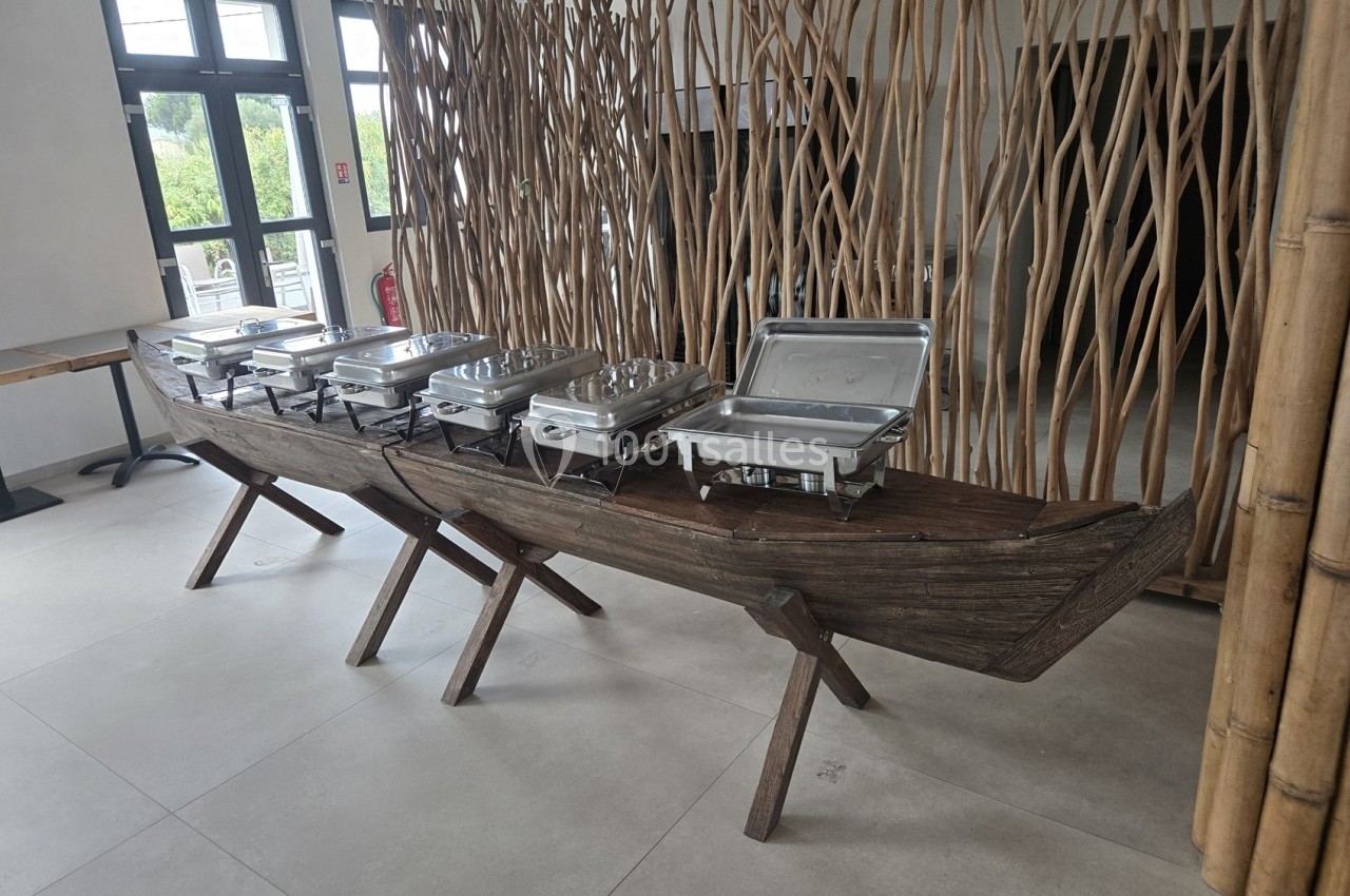 Buffet présenté sur une longue pirogue en bois avec des plats en inox, dans une salle lumineuse au décor naturel.