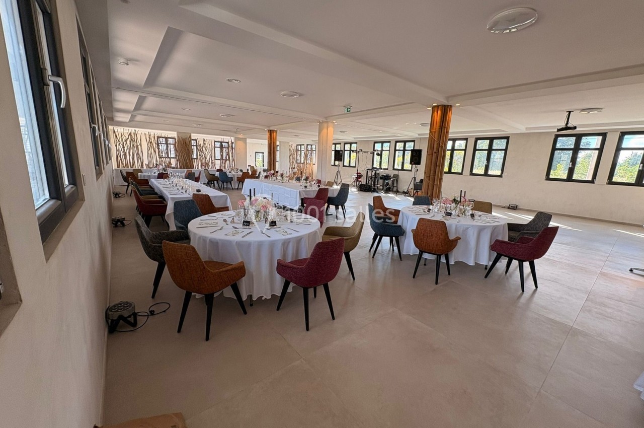 Salle lumineuse avec tables rondes dressées, nappes blanches et chaises colorées, prête pour un événement.