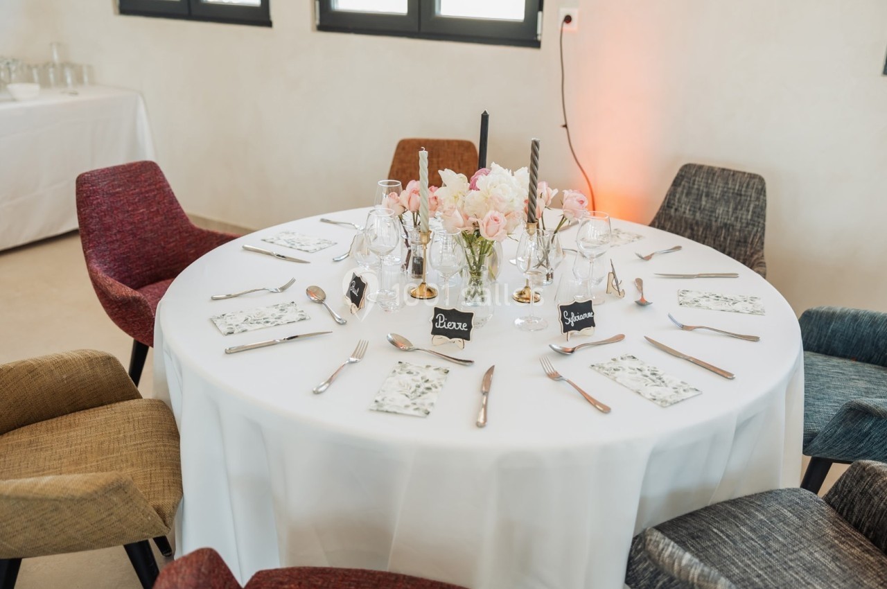 Table ronde dressée pour un repas, avec nappes blanches, couverts, verres, fleurs et marque-places.