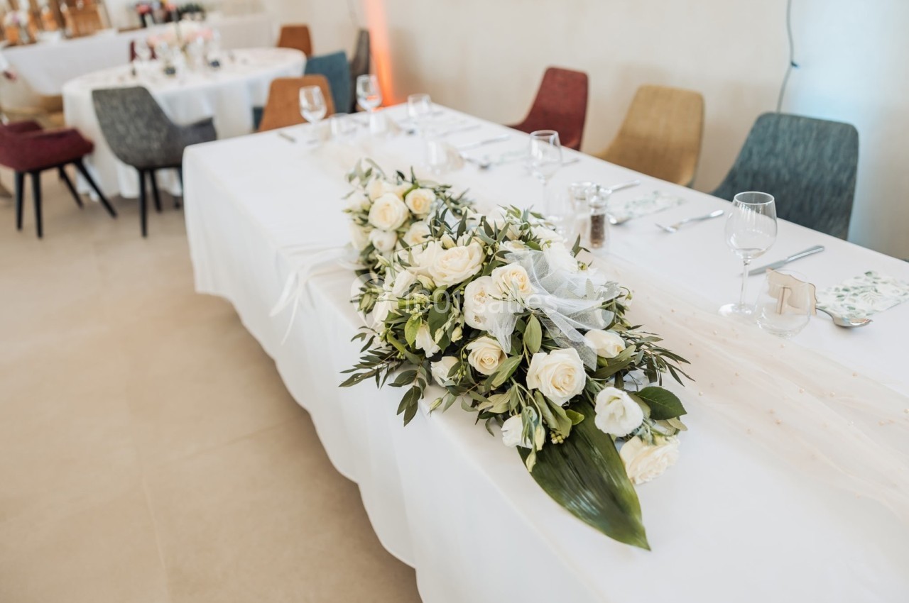 Centre de table avec des roses blanches et feuillage, disposé sur une nappe blanche dans une salle de réception.