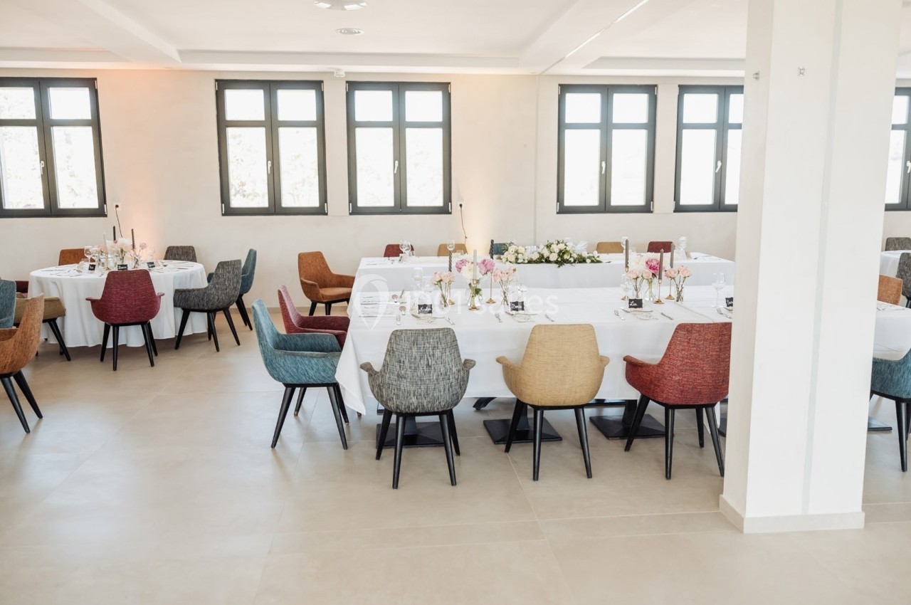 Salle lumineuse avec tables dressées, nappes blanches, chaises colorées et décoration florale.