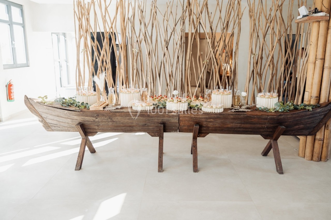 Table en forme de barque en bois, décorée de plantes et de desserts, placée dans un espace lumineux.