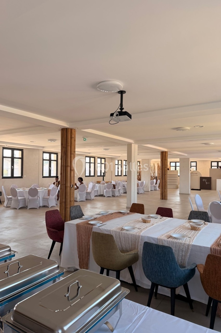 Salle lumineuse aménagée pour un événement, avec tables dressées, chaises colorées et buffets en premier plan.