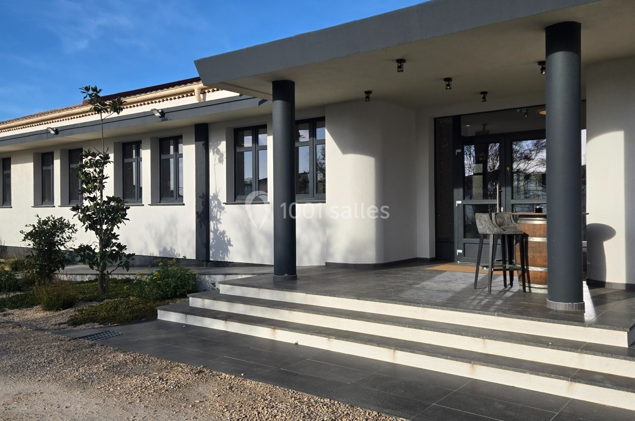Façade d'un bâtiment moderne avec terrasse, colonnes noires et entrée vitrée, entouré de végétation.