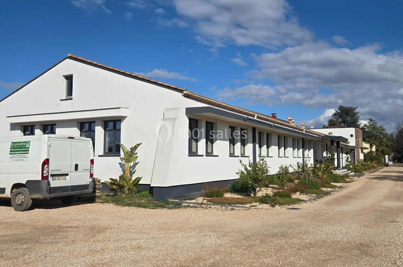 Bâtiment blanc de plain-pied avec des fenêtres noires, une allée en gravier et un utilitaire stationné à gauche.