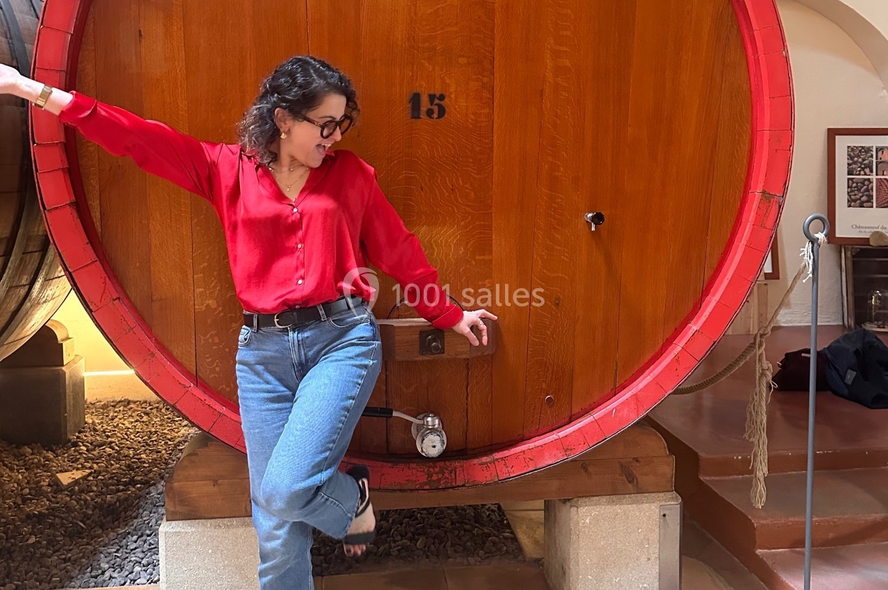 Femme en chemise rouge posant devant un grand tonneau en bois dans un environnement intérieur chaleureux.