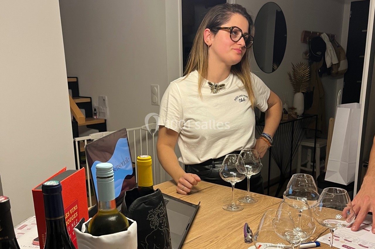 Une femme debout près d'une table avec des verres à vin, des bouteilles et divers objets dans un intérieur moderne.