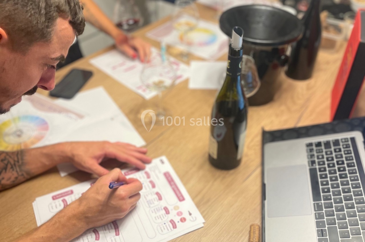 Un homme remplit un document sur le vin à une table avec une bouteille, un ordinateur et des fiches de dégustation.