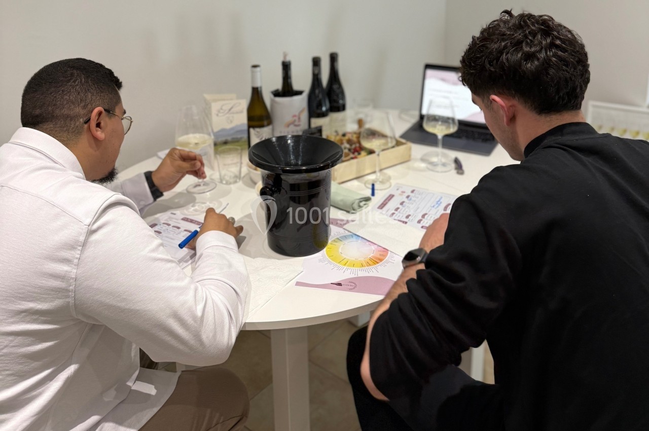 Deux hommes assis à une table dégustent du vin tout en prenant des notes, avec des bouteilles et un ordinateur en arrière…