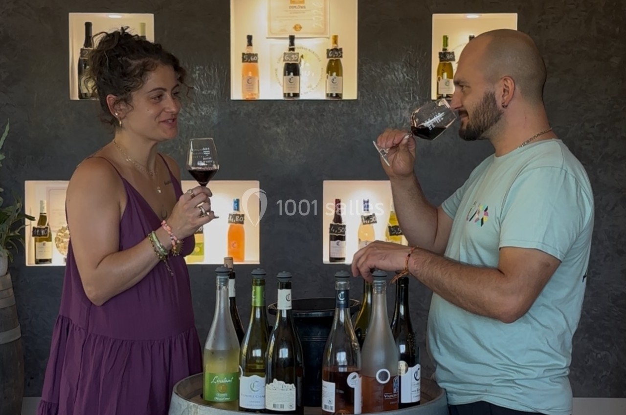 Deux personnes dégustent du vin devant un présentoir de bouteilles éclairé, avec un tonneau en premier plan.