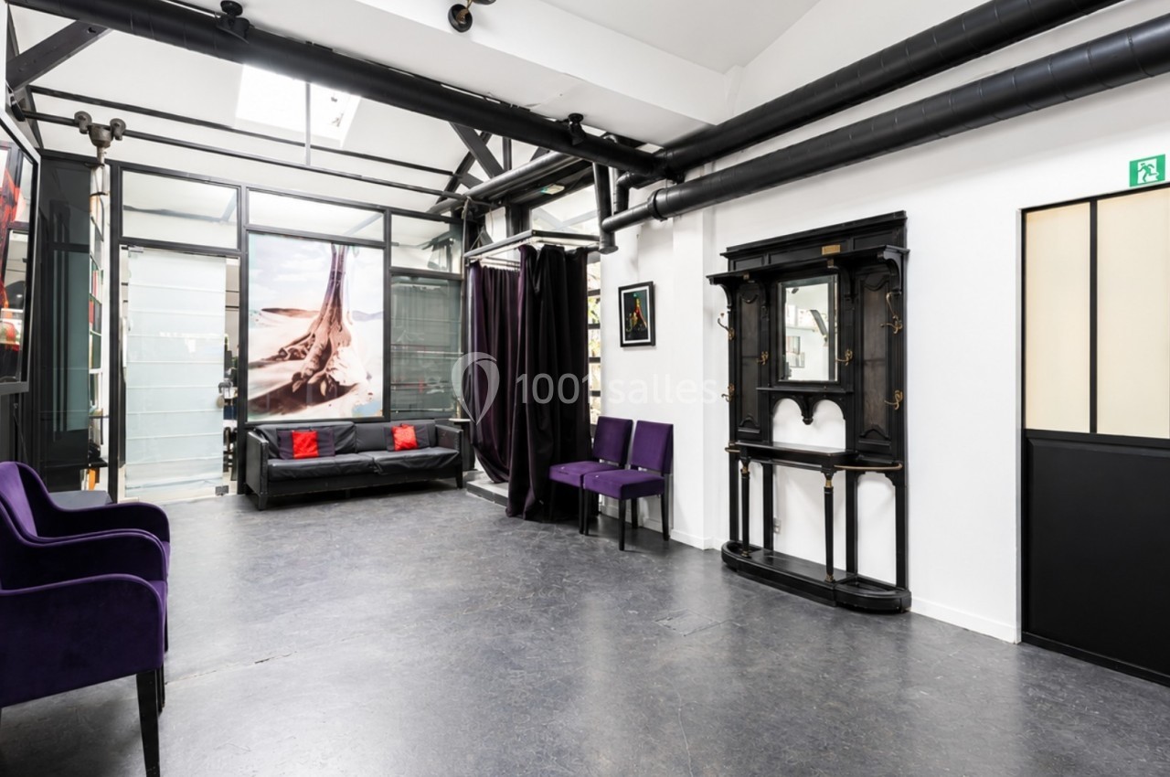 Salle lumineuse avec mobilier violet, miroir mural, canapé noir et grandes baies vitrées donnant sur un espace intérieur.