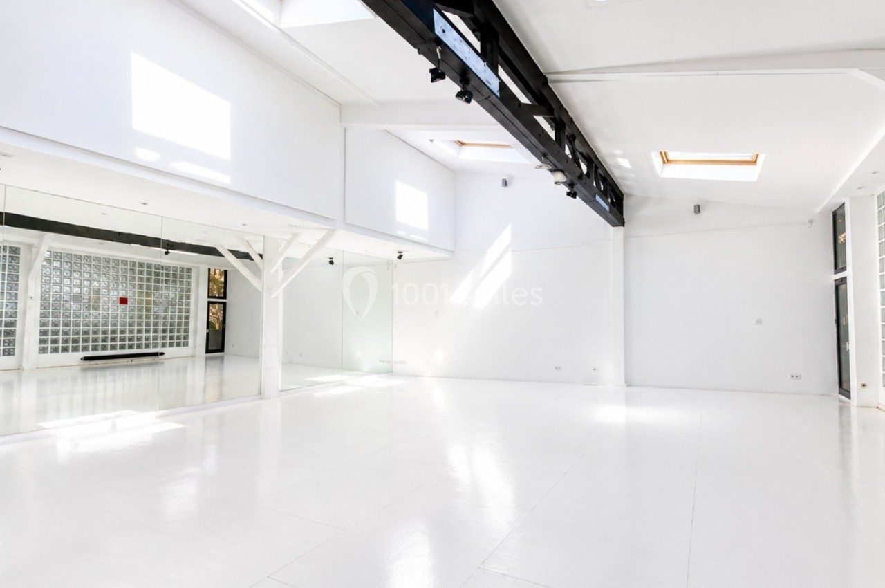 Salle lumineuse avec murs blancs, sol en parquet clair, grandes fenêtres et miroirs, idéale pour activités intérieures.