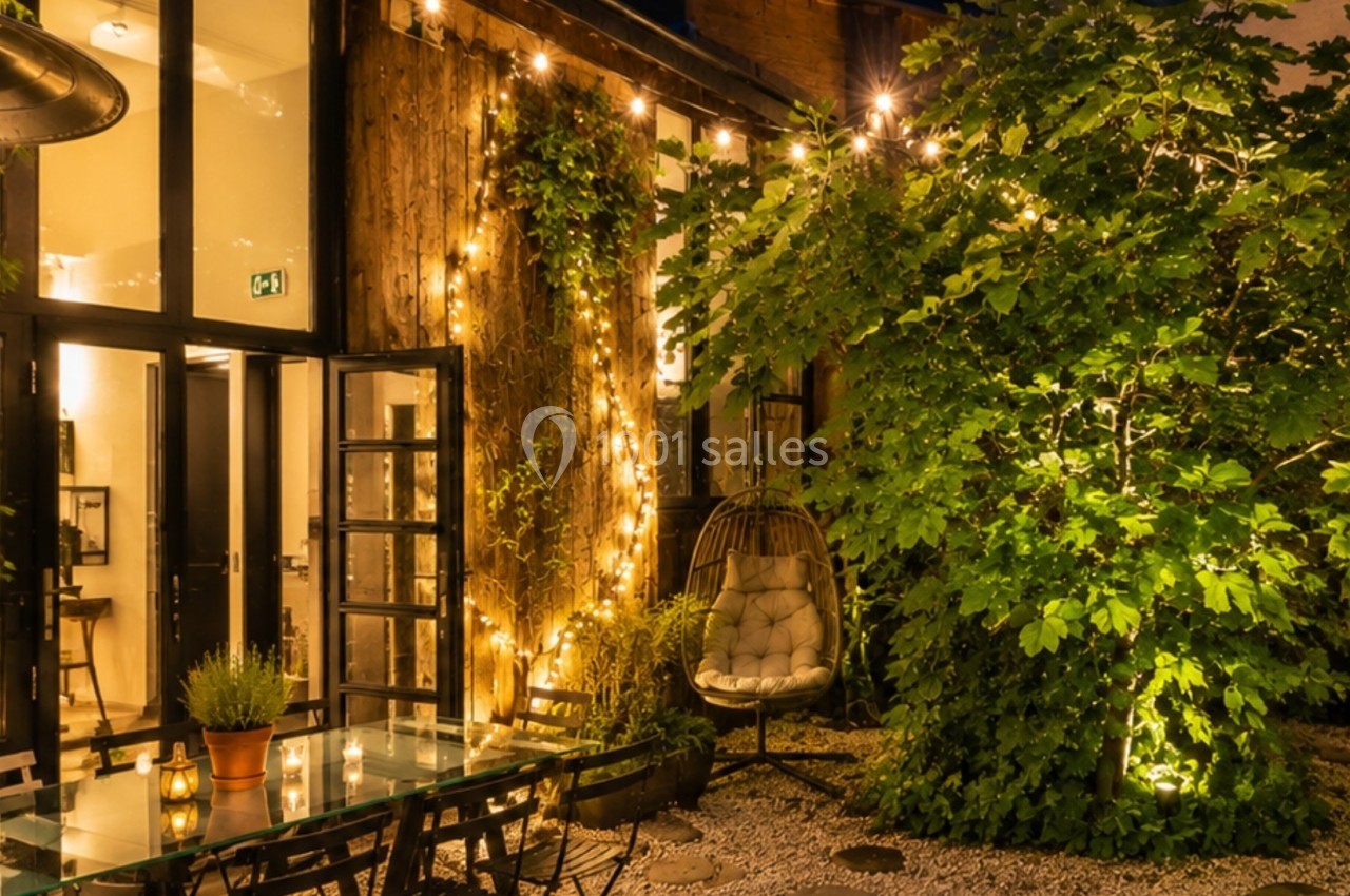 Terrasse extérieure éclairée par des guirlandes lumineuses, avec table en verre, plantes et fauteuil suspendu.