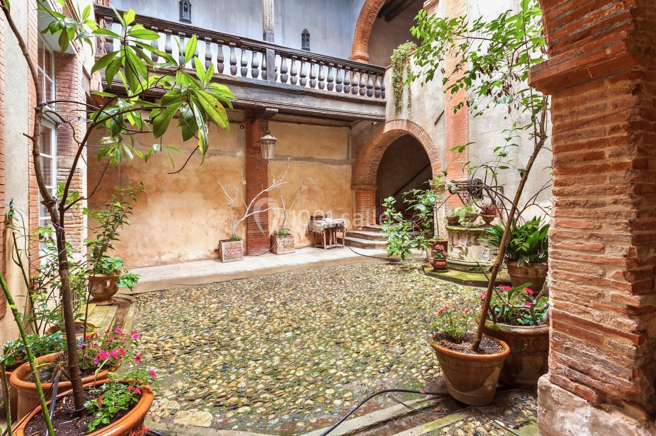 Cour intérieure pavée avec des plantes en pots, entourée de murs en briques et d'arches, éclairée naturellement.