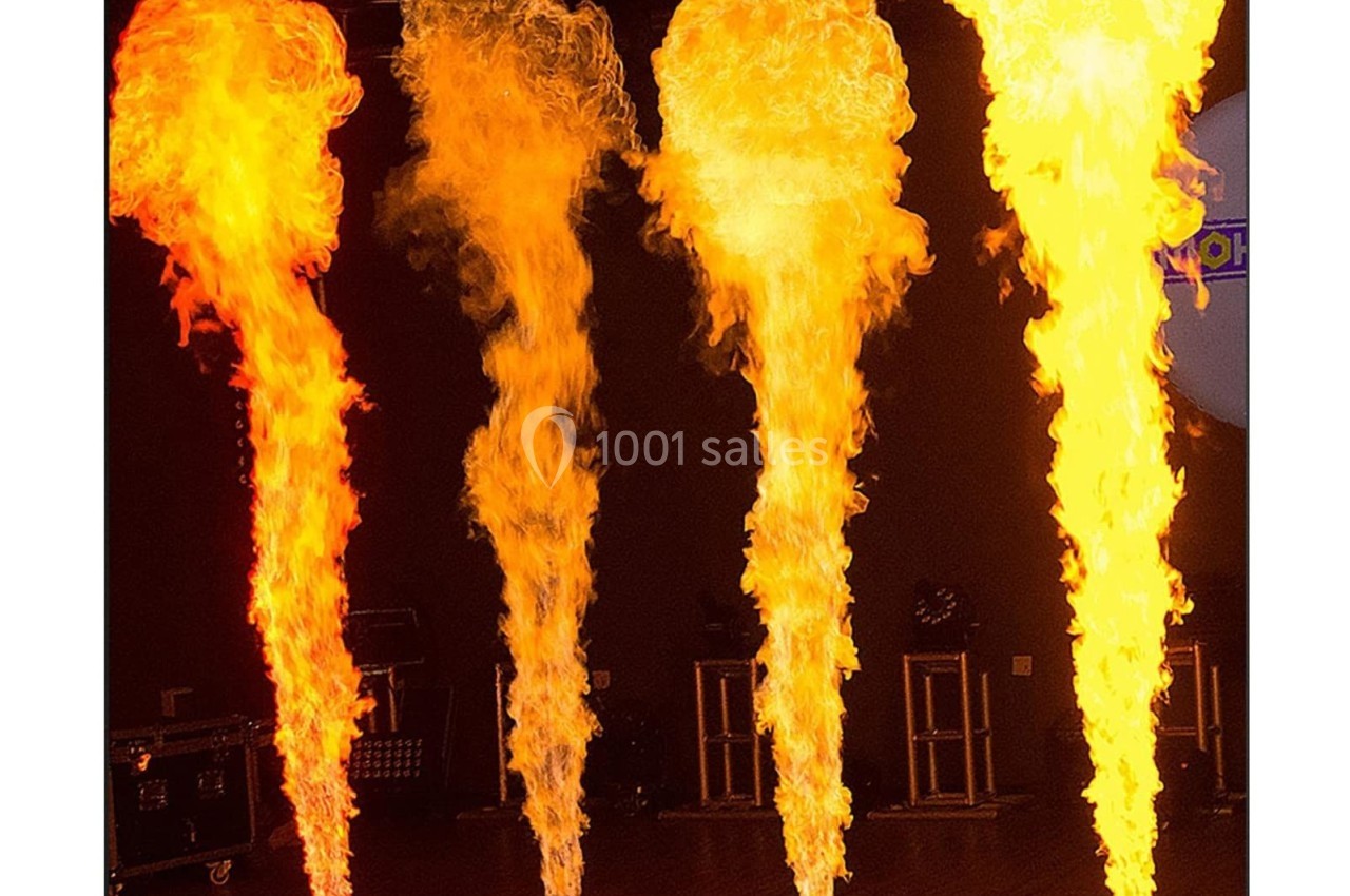 Quatre colonnes de flammes jaillissent de dispositifs au sol dans une salle sombre.