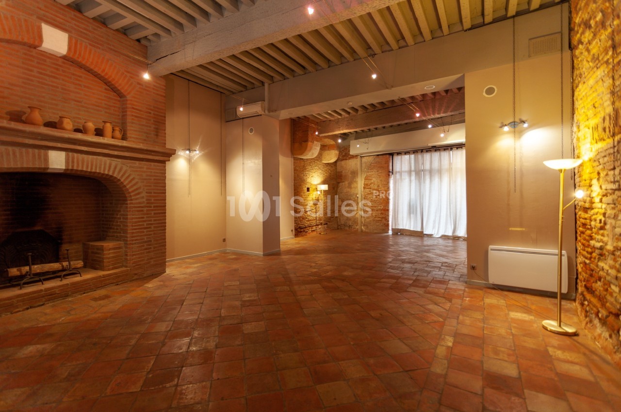 Salle vide avec sol en terre cuite, murs en briques, cheminée, éclairage tamisé et grande baie vitrée avec rideaux.