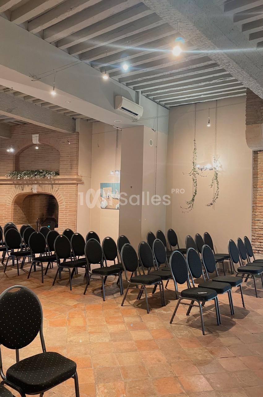 Salle avec des rangées de chaises noires alignées, sol en terre cuite, murs en briques et plafond avec poutres apparentes.