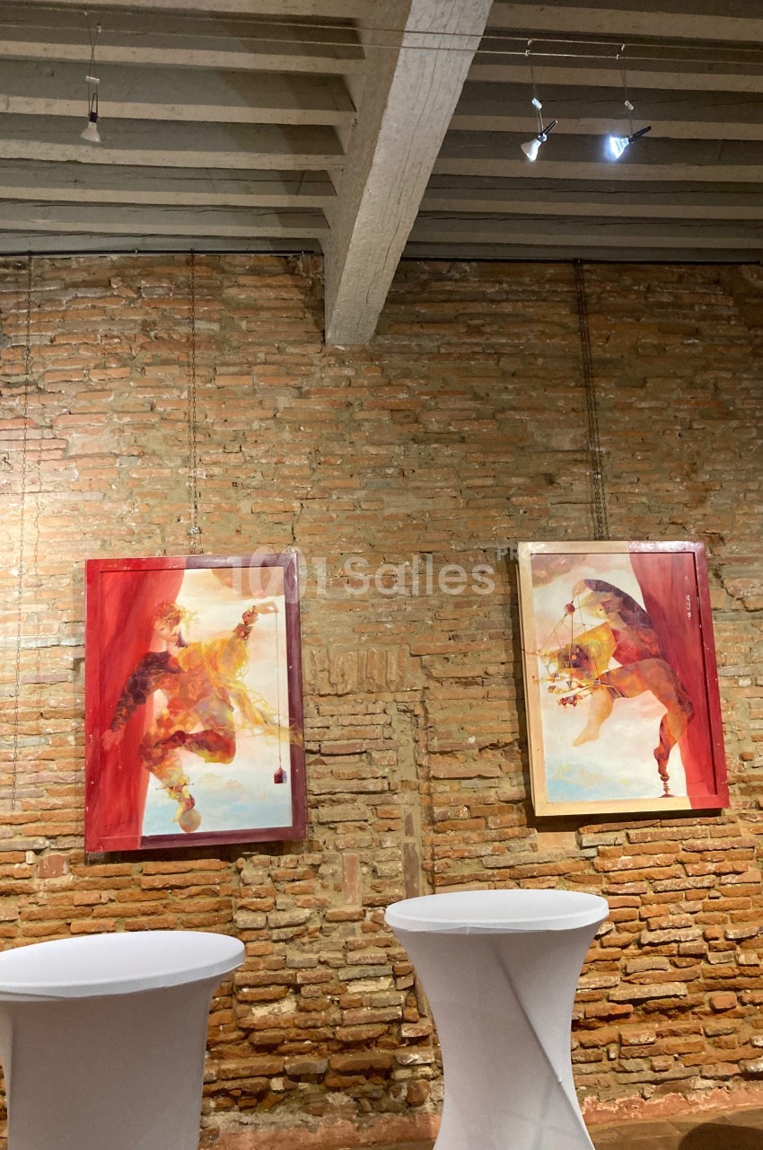 Deux tableaux colorés accrochés sur un mur en briques, avec des tables hautes blanches au premier plan.