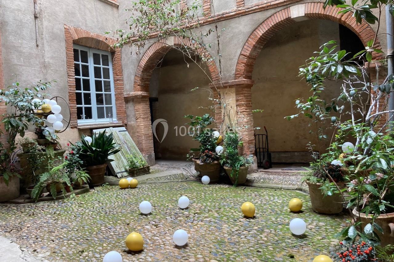 Cour intérieure pavée avec des arches en briques, des plantes en pots et des ballons blancs et dorés dispersés au sol.