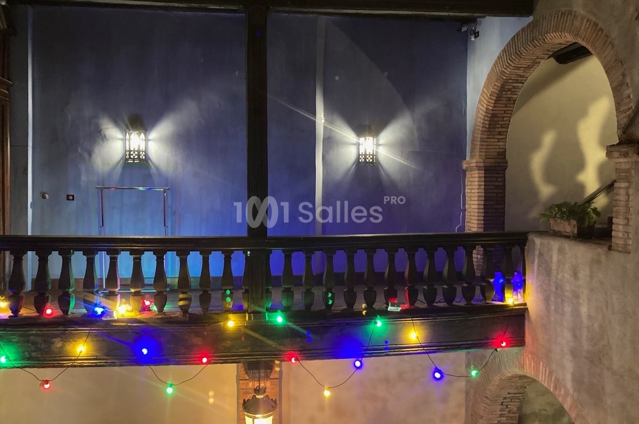 Balustrade en bois décorée de guirlandes lumineuses colorées, éclairée par des appliques murales sur un mur bleu.