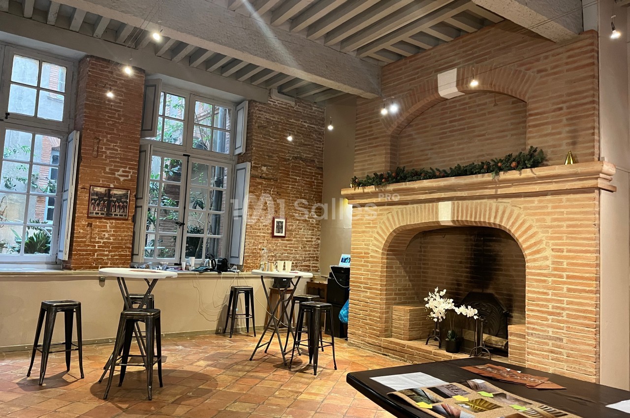 Salle lumineuse avec murs en briques, cheminée décorée, tables hautes et tabourets, sol en carreaux de terre cuite.