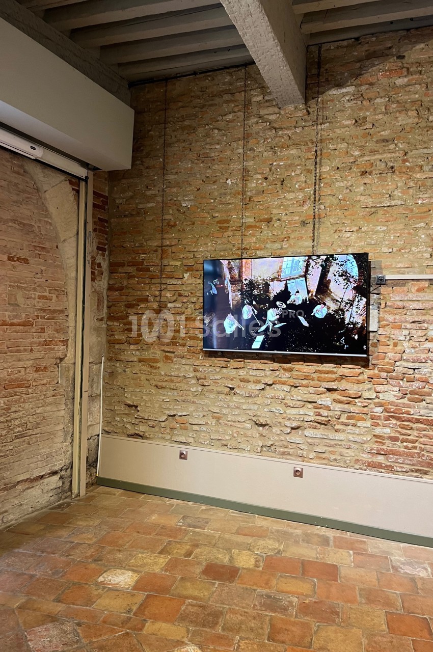 Mur en briques apparentes avec un écran fixé affichant une vidéo, sol en carrelage et plafond avec poutres en bois.