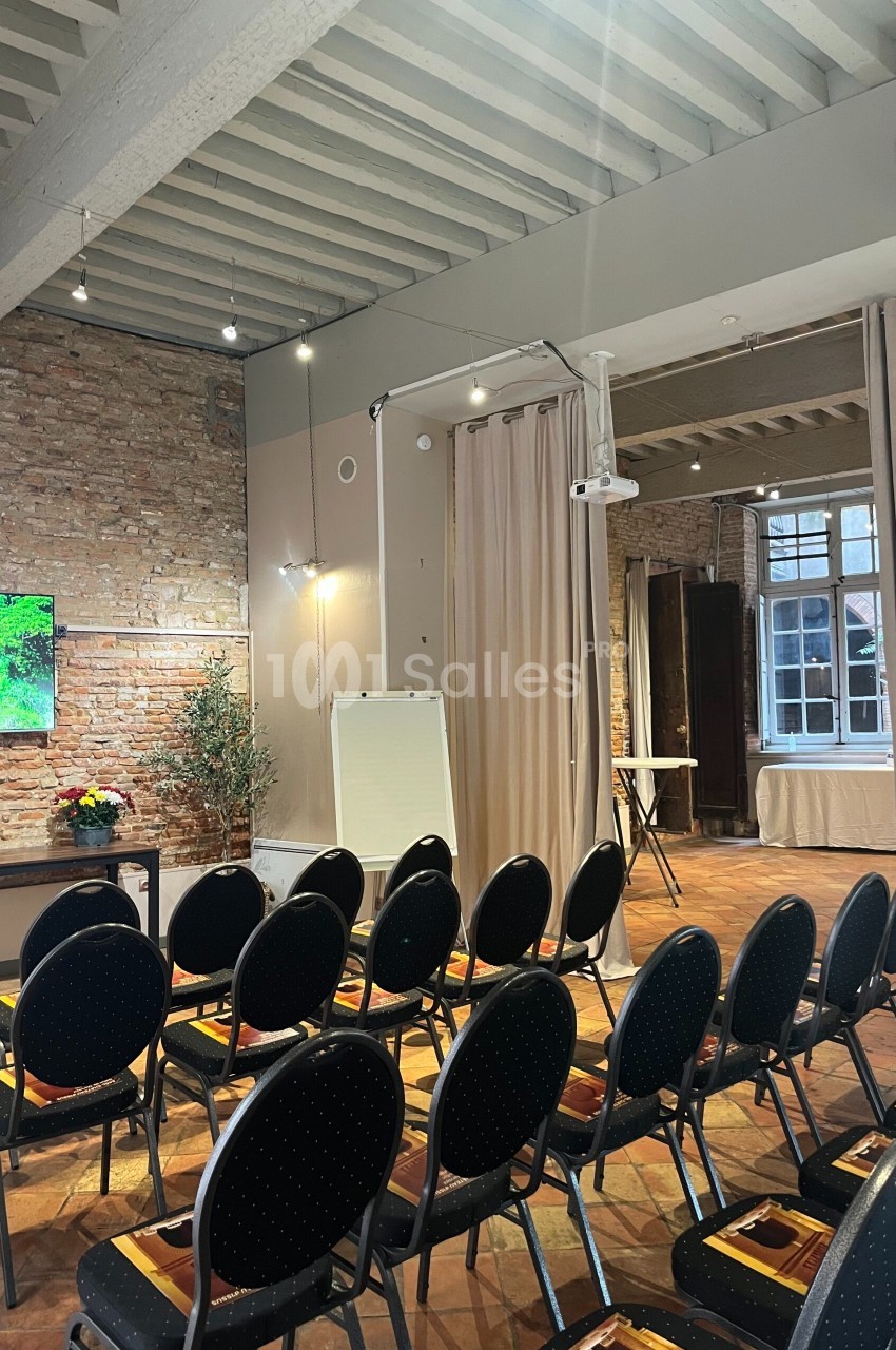 Salle de réunion avec chaises alignées, tableau blanc, écran mural, murs en briques et lumière naturelle.