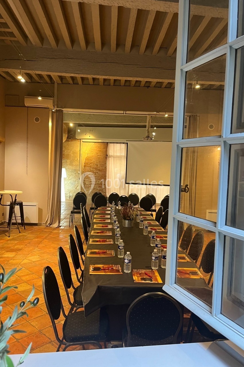 Salle de réunion avec une grande table rectangulaire dressée, entourée de chaises noires, éclairée par une lumière naturelle.