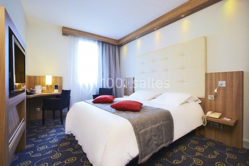 Chambre d'hôtel lumineuse avec un grand lit, deux coussins rouges, bureau, télévision et fenêtre avec rideaux.