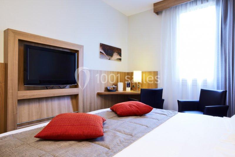 Chambre lumineuse avec lit double, coussins rouges, télévision murale, bureau, lampe et fauteuils près d'une fenêtre.