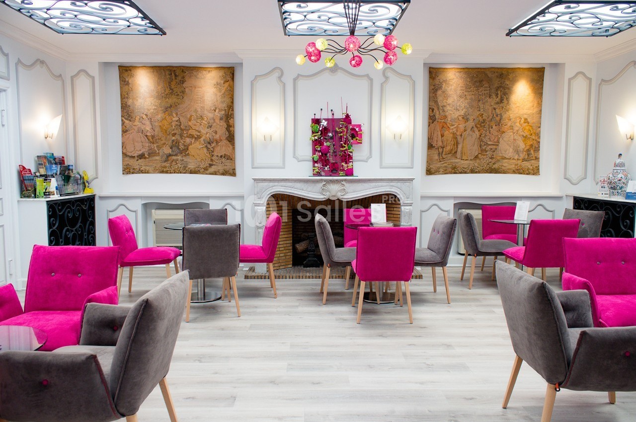 Salle lumineuse avec des fauteuils roses et gris, tables en verre, cheminée décorative et tapisseries murales.