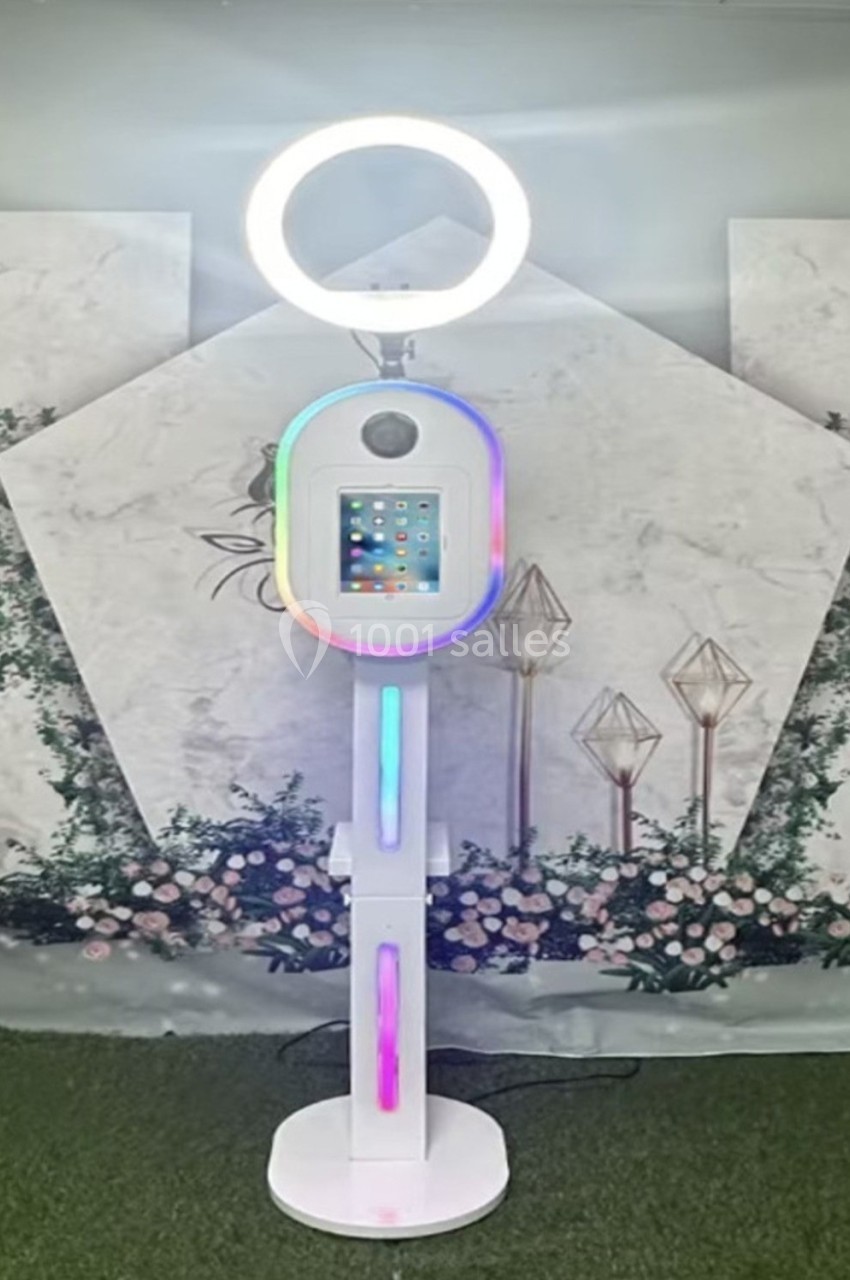 Borne photo interactive avec écran tactile et anneau lumineux, placée devant un décor floral et géométrique.