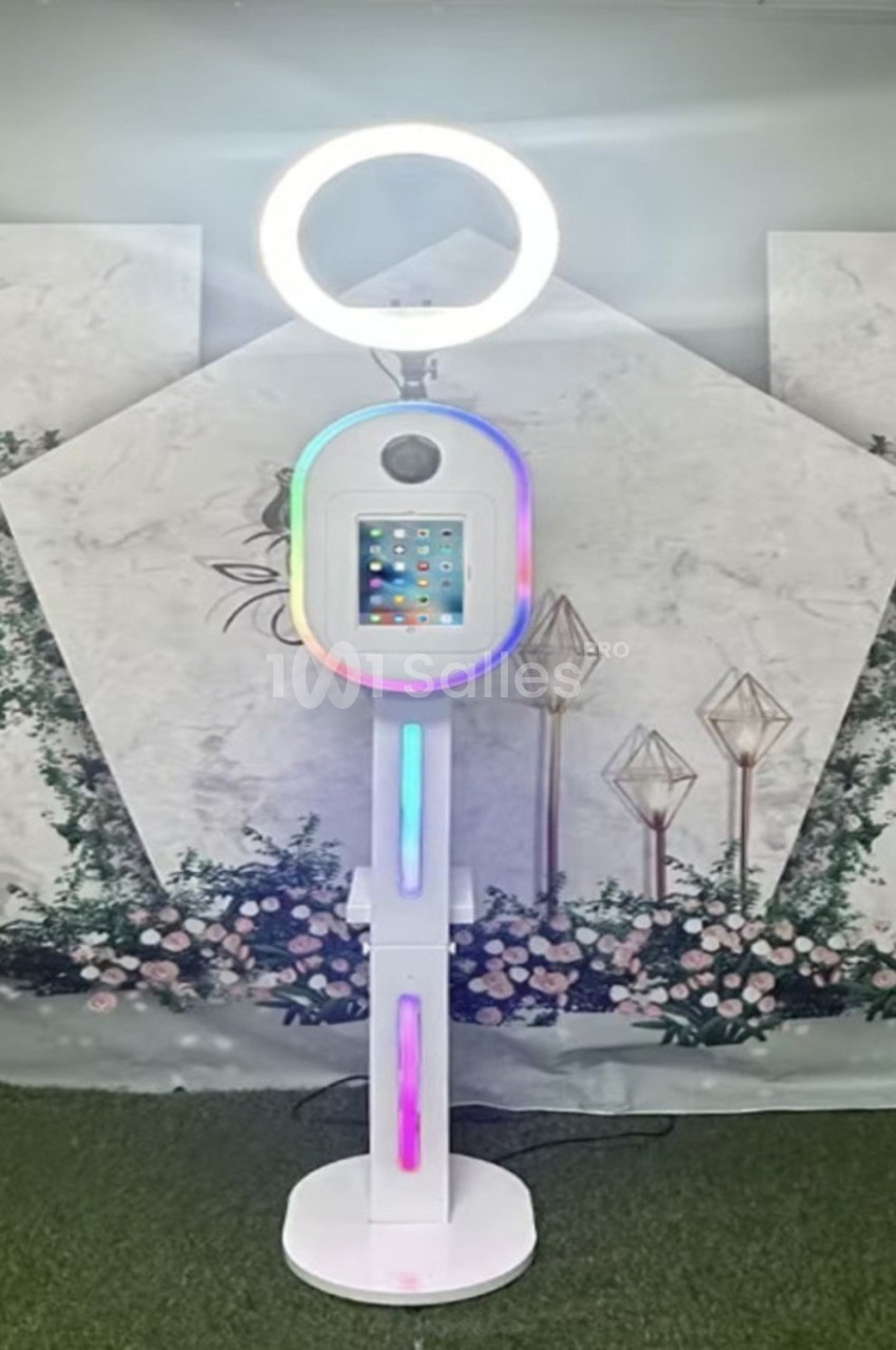 Borne photo interactive avec écran tactile et anneau lumineux, placée devant un décor floral et géométrique.