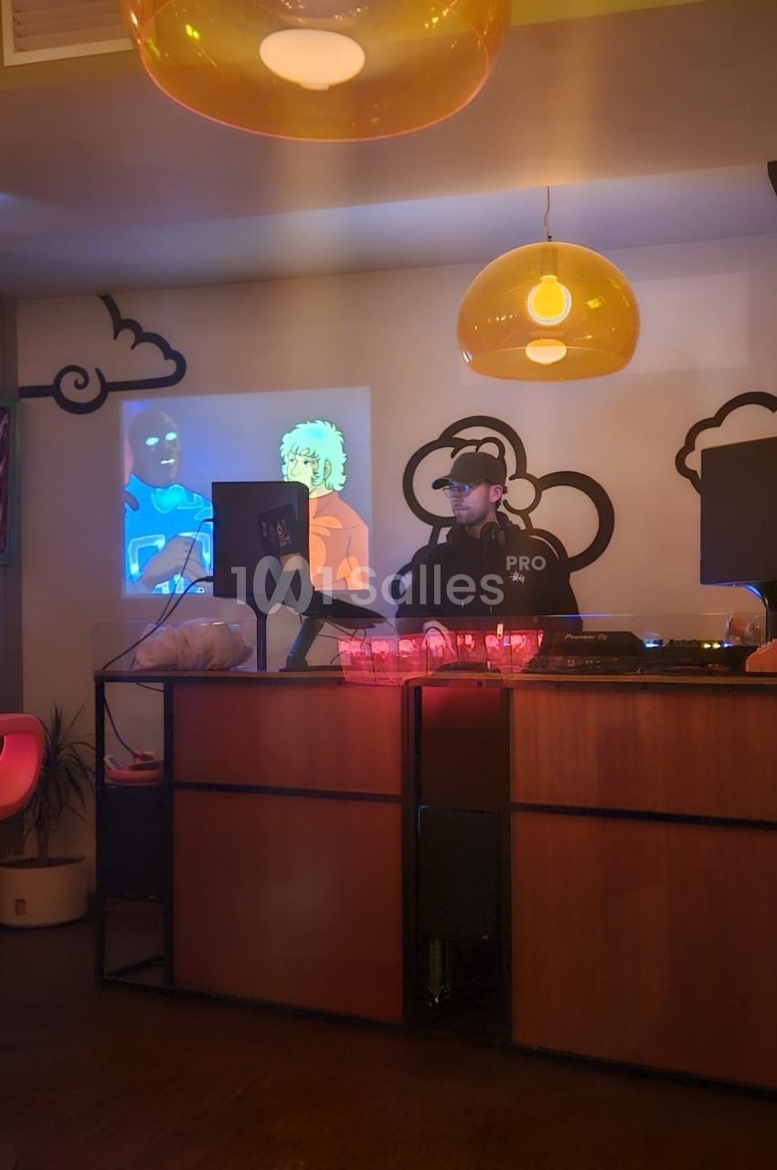 DJ en cabine derrière un pupitre bois, équipement audio visible, écrans d'ambiance colorés, luminaires suspendus dorés, décoration nuages stylisés.