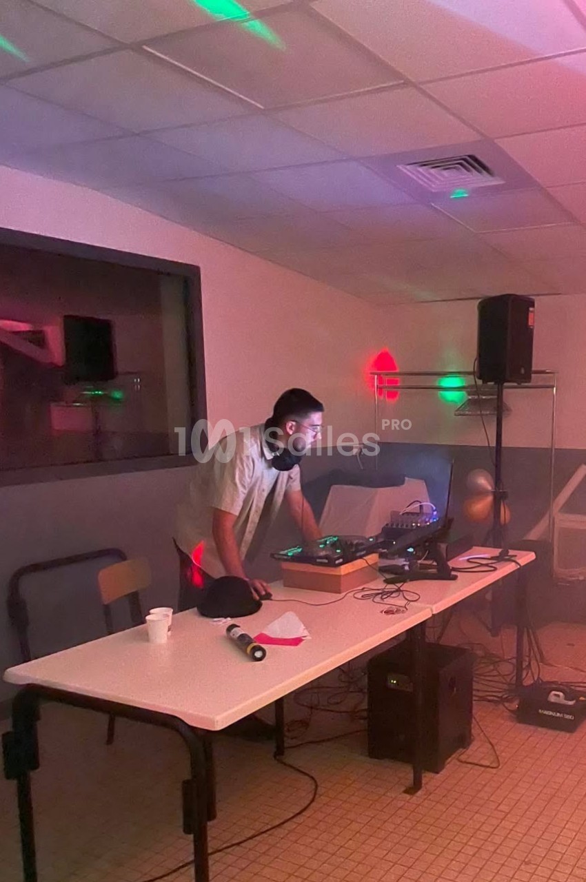 DJ en stand en intérieur face à sa console, éclairage rose et vert, enceintes et équipement autour.