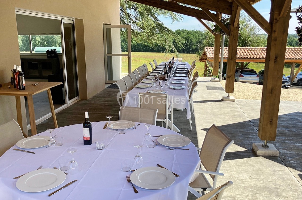Tables dressées avec nappes blanches, couverts et verres, sur une terrasse couverte avec vue sur un paysage champêtre.