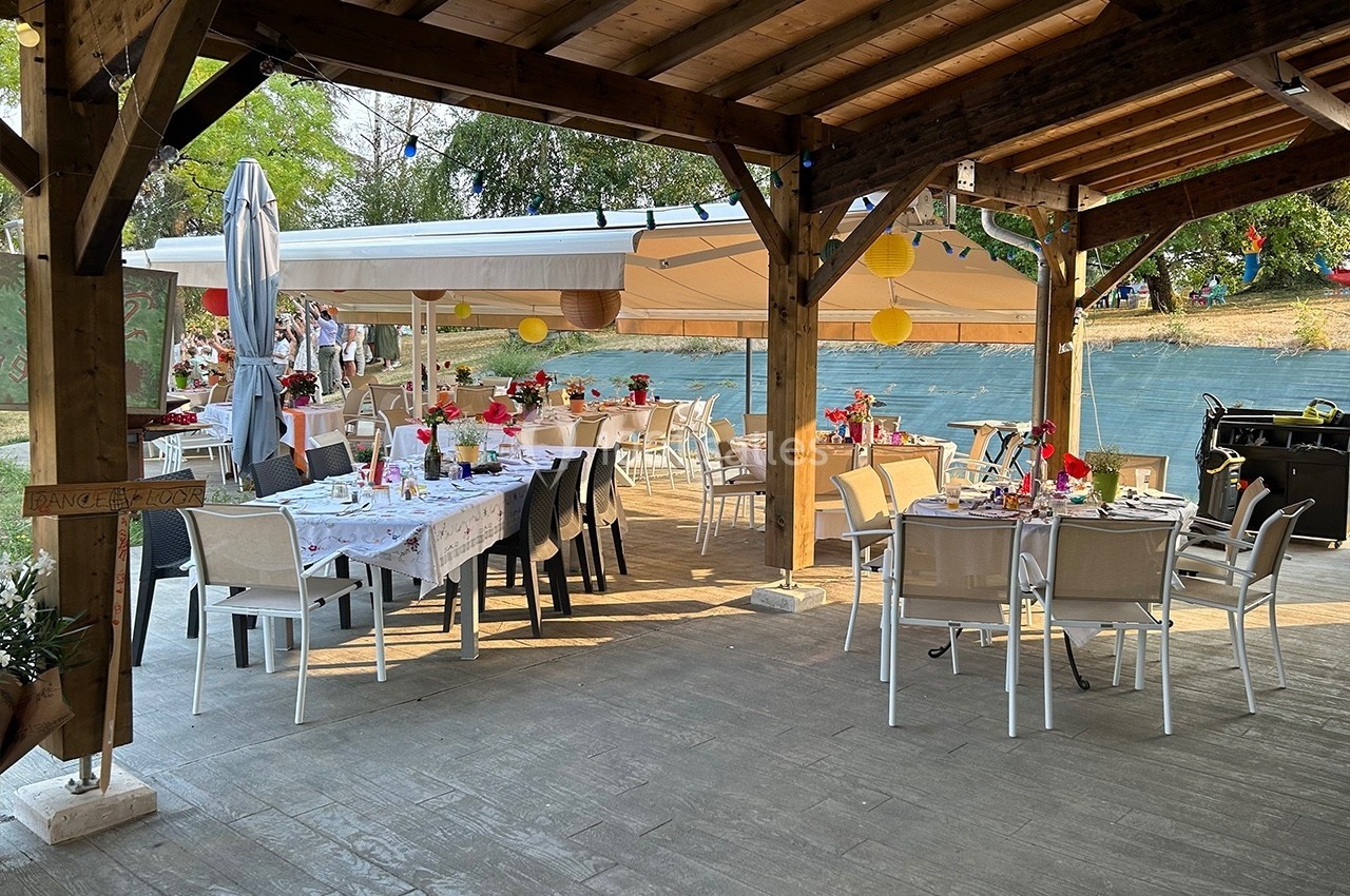 Terrasse couverte aménagée pour un repas, avec tables décorées et vue sur un plan d'eau en arrière-plan.